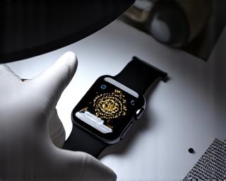 Ruka v bílé rukavici drží Apple Watch a kontroluje kvalitu gravírování lupou, pro finální kontrolu.